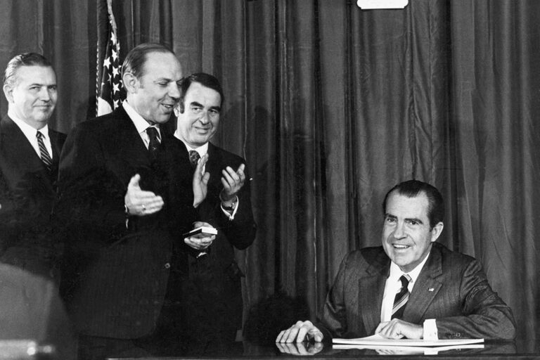 President Richard Nixon signs sitting down at a desk with the Occupational Health and Safety Act signed in front of him. Three government officials smile and stand off to the left.