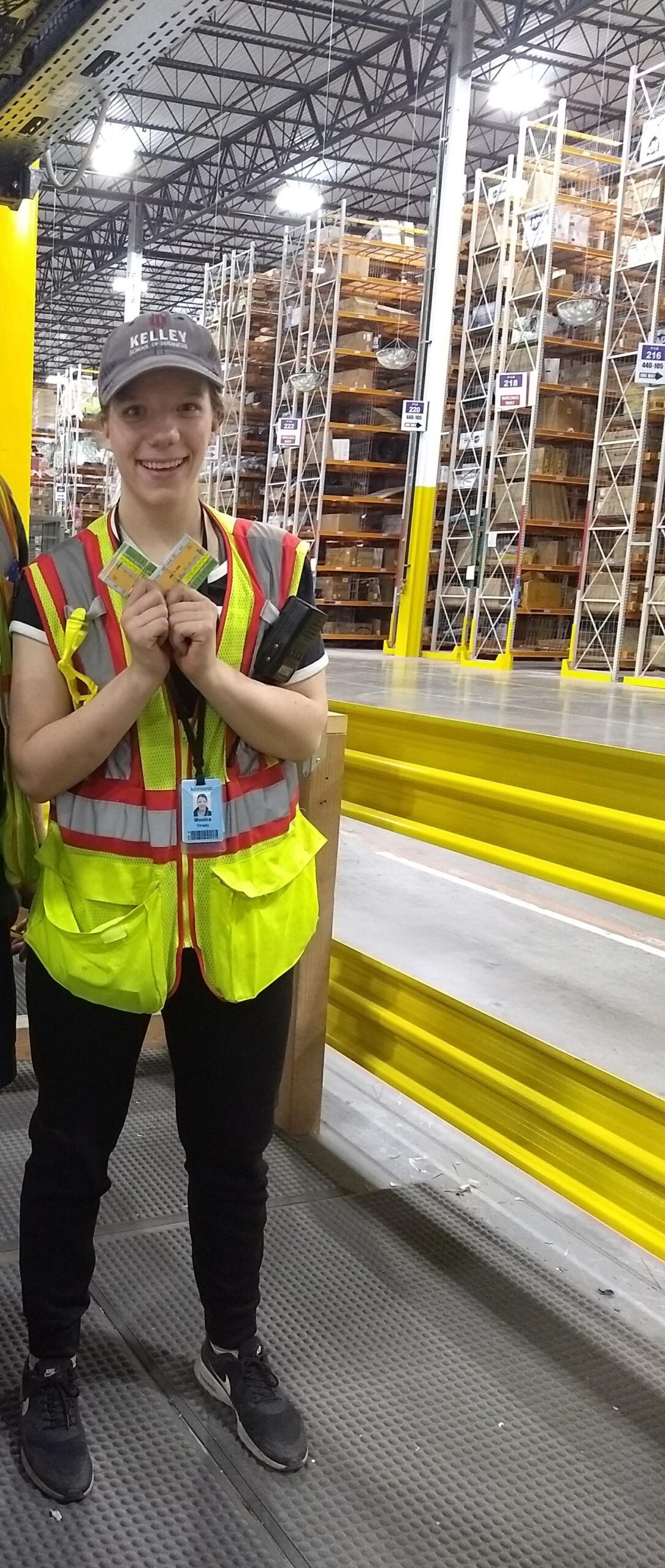 Female warehouse worker smiling in an oversized high visibility vest with big warehouse in the background.